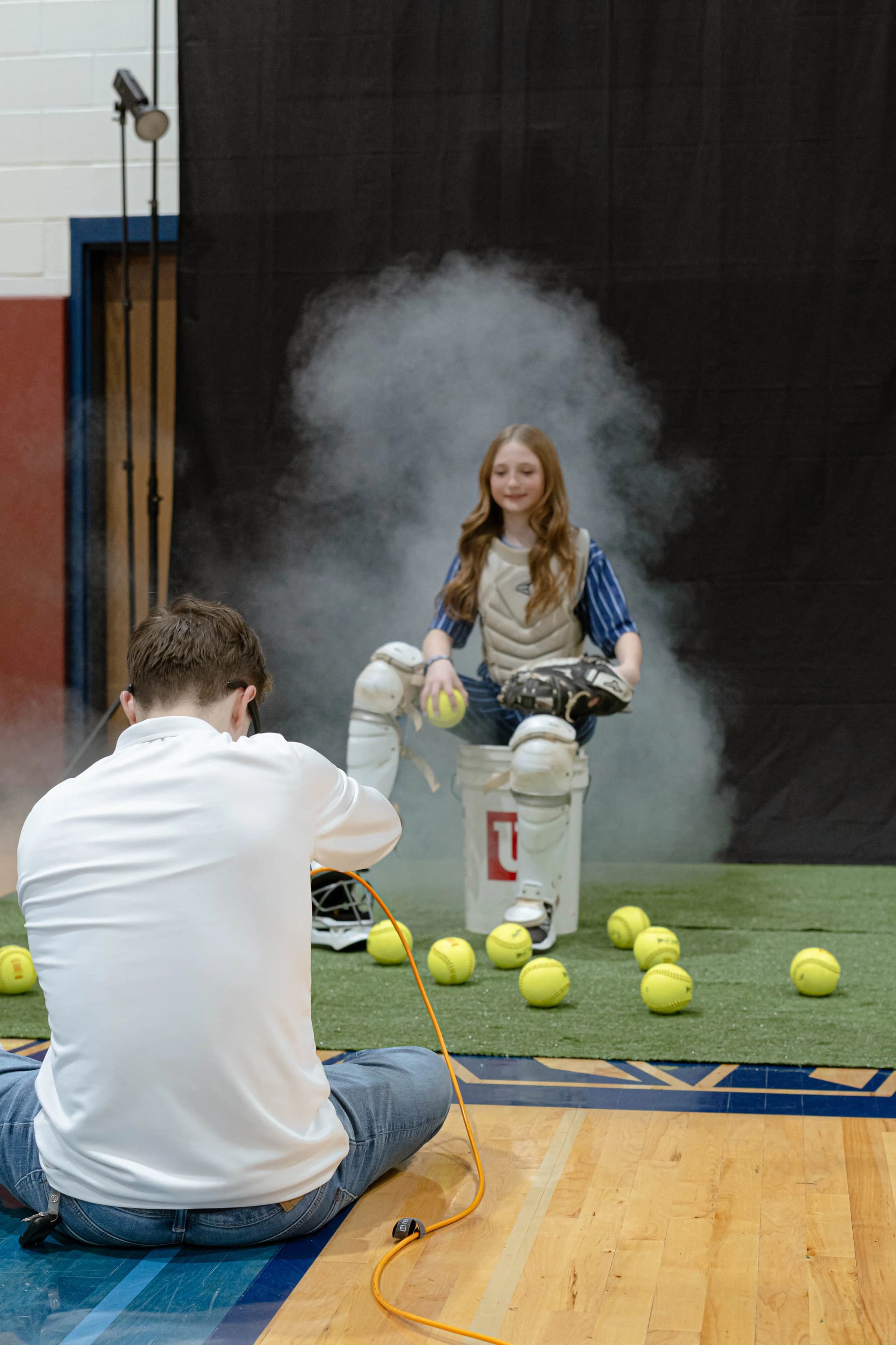 Cole photographing a softball catcher posed on a ball bucket as smoke rises behind her