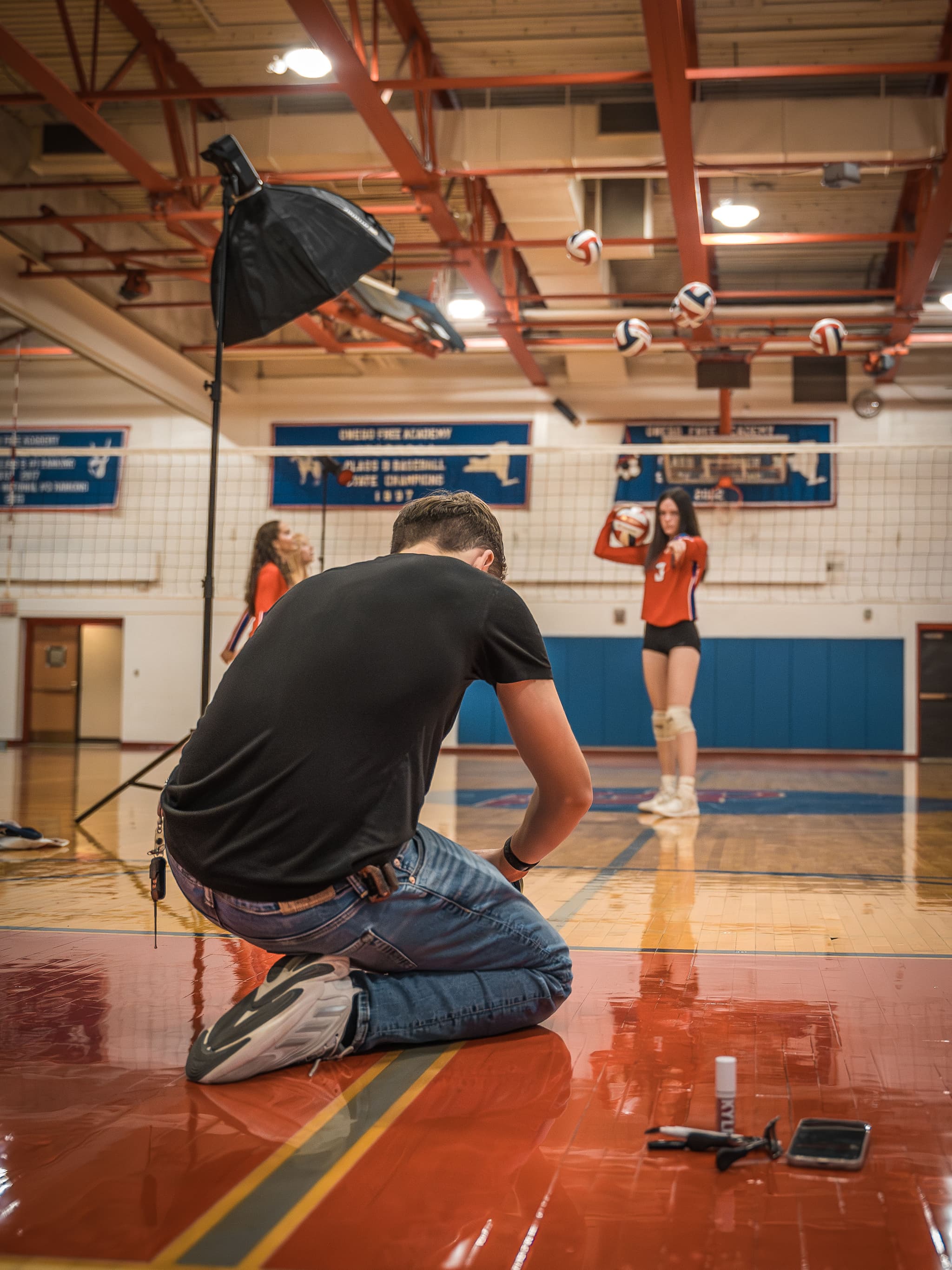 Cole kneeling behind the camera with a softbox raised, volleyballs suspended in the air and an athlete waiting on the far side of the net